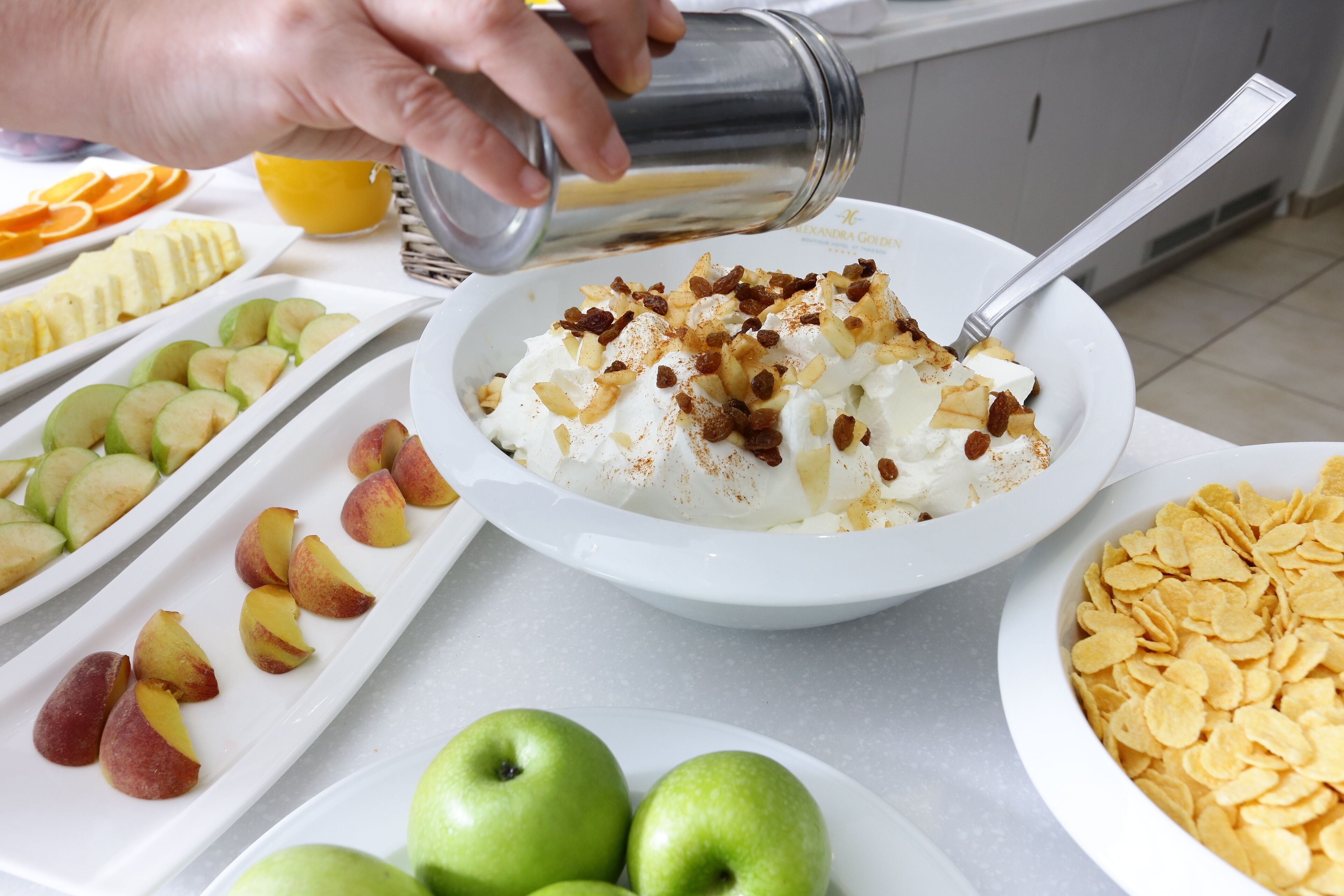 A breakfast setting with a bowl of cereal topped with whipped cream and nuts, alongside plates of sliced fruits and cereal flakes.