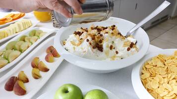 A breakfast setting with a bowl of cereal topped with whipped cream and nuts, alongside plates of sliced fruits and cereal flakes.