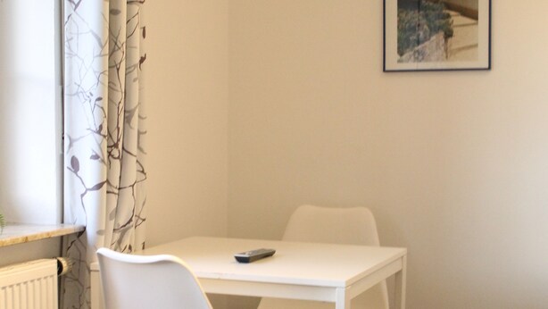 Une petite pièce propre, avec une table blanche et deux chaises blanches, un radiateur et un tableau encadré au mur.