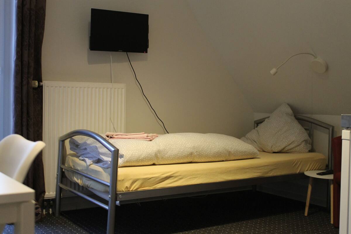 Una cama individual con cabecera, una mesita de noche, una silla, un televisor de pantalla plana montado en la pared y una ventana con cortinas.