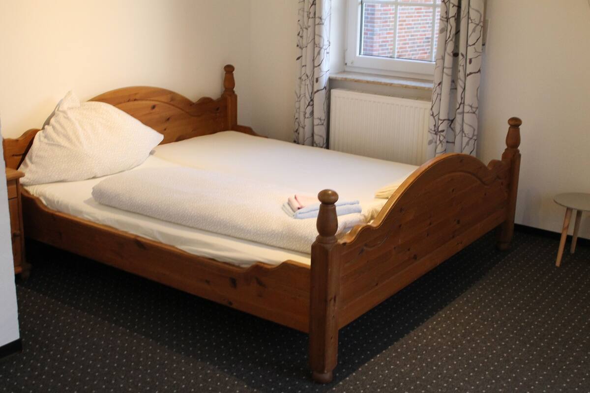 Una cama de madera individual con colchón y almohadas blancas, una mesita de noche pequeña y una ventana con cortinas estampadas.