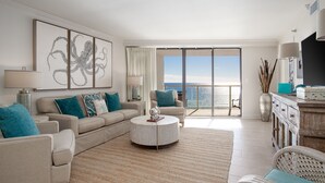 Una sala de estar moderna con un sofá color beige, cojines verde azulado, una mesa de centro redonda y un ventanal con vista al océano.