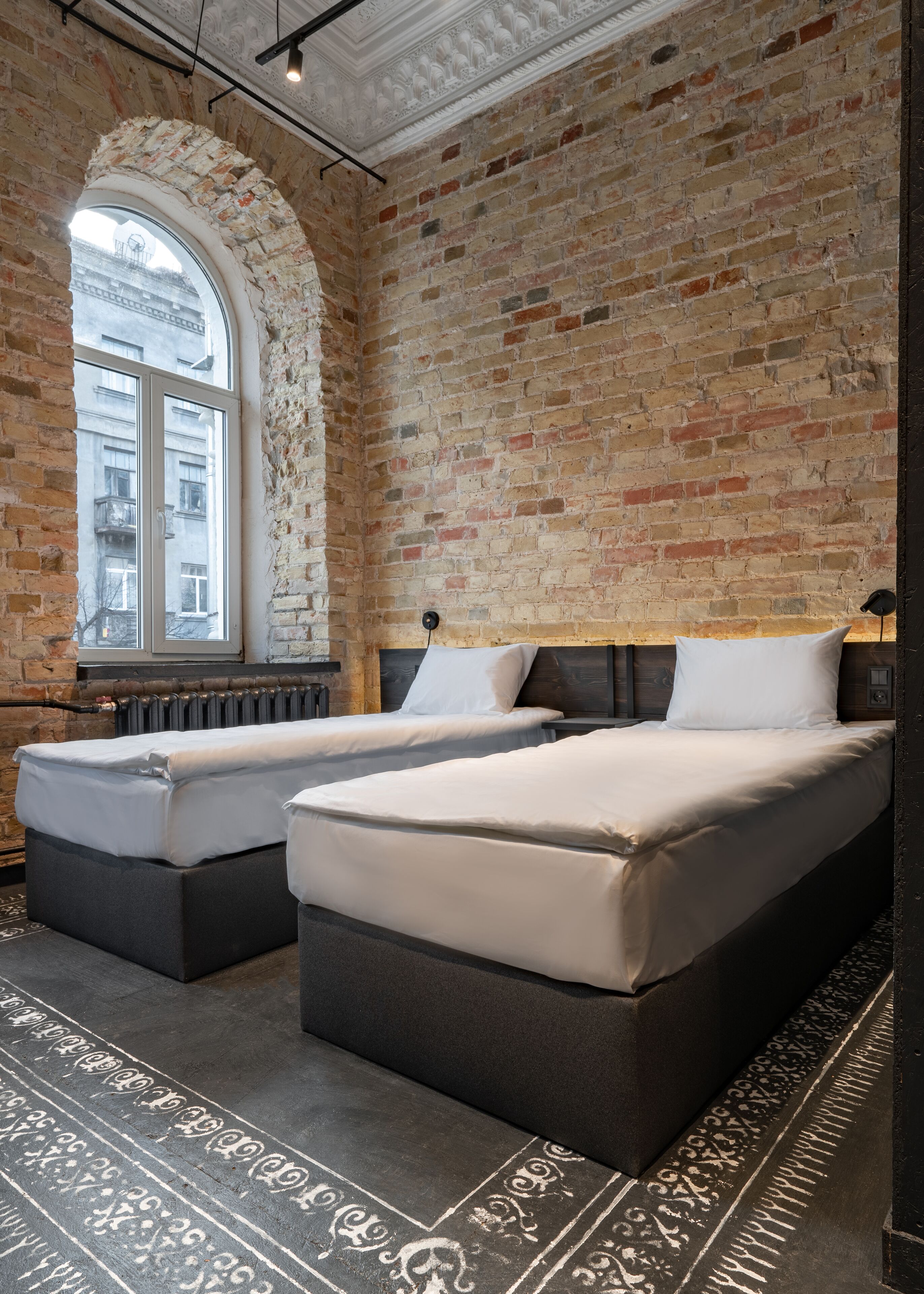 A room with two beds, a brick wall, a large window, and a radiator.