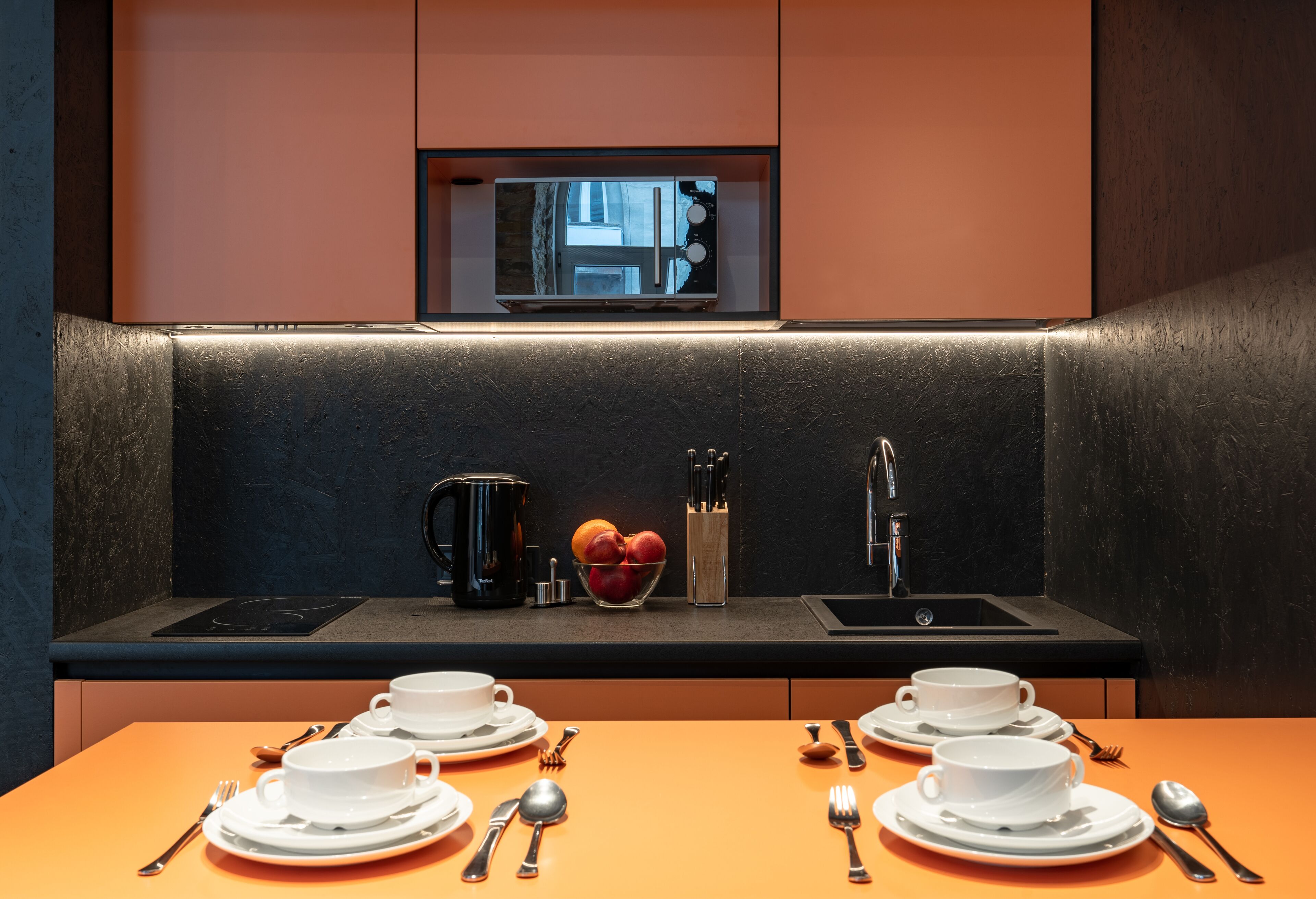 A modern kitchen with a built-in microwave, a sink, and a countertop with a bowl of fruit and utensils.