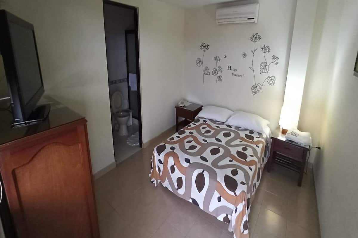 A bedroom with a bed, a desk, a chair, a television, and a bathroom visible through an open door.