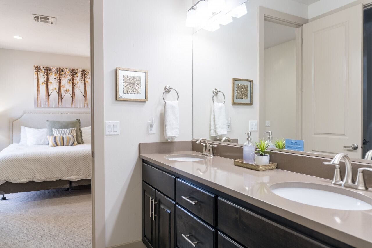 A modern bathroom with two sinks, a large mirror, and a bed with a view of the room.