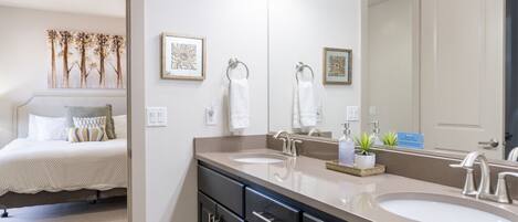 A modern bathroom with two sinks, a large mirror, and a bed with a view of the room.