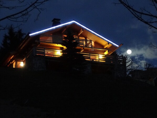 A wooden cabin with a balcony, illuminated by warm interior lighting, set against a night sky with a visible moon. - Luxury Chalet with Ski-In/Ski-Out, Sauna & Outdoor Jacuzzi at Le Grand-Bornand (Le Grand-Bornand)
