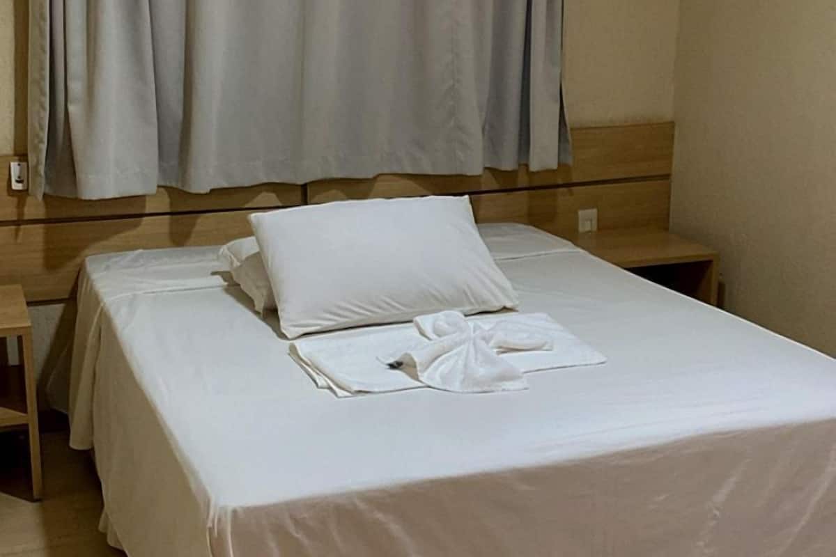 A neatly made bed with white linens and pillows, a wooden nightstand, a small table, and a wall-mounted air conditioner.