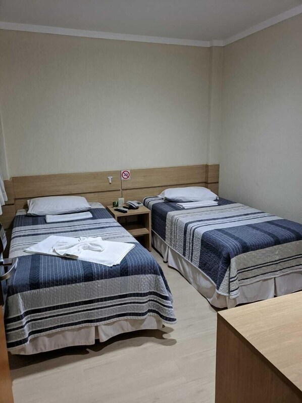 A hotel room with two beds, each with striped bedding and pillows. There is a wooden headboard, a bedside table with a phone, and a no-smoking sign on the wall. - Onix Inn Hotel (Teodoro Sampaio)