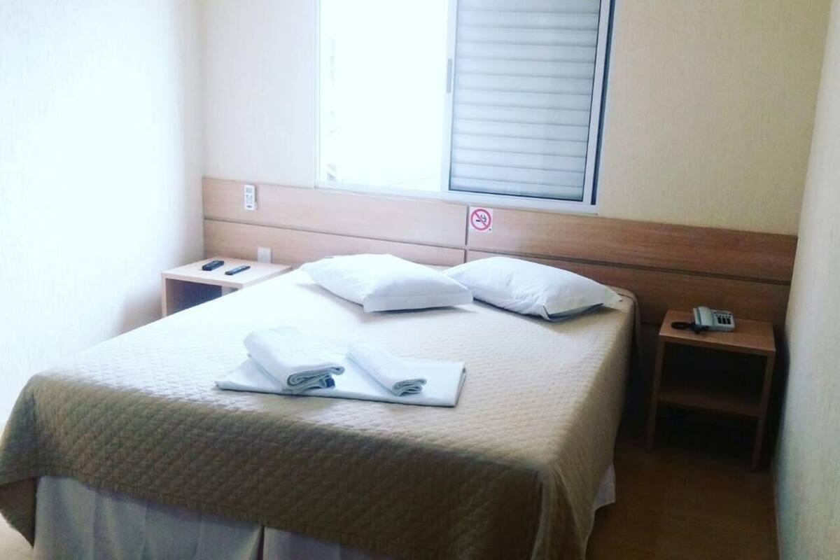 A hotel room with a bed, a bedside table with a phone, and a window with blinds.