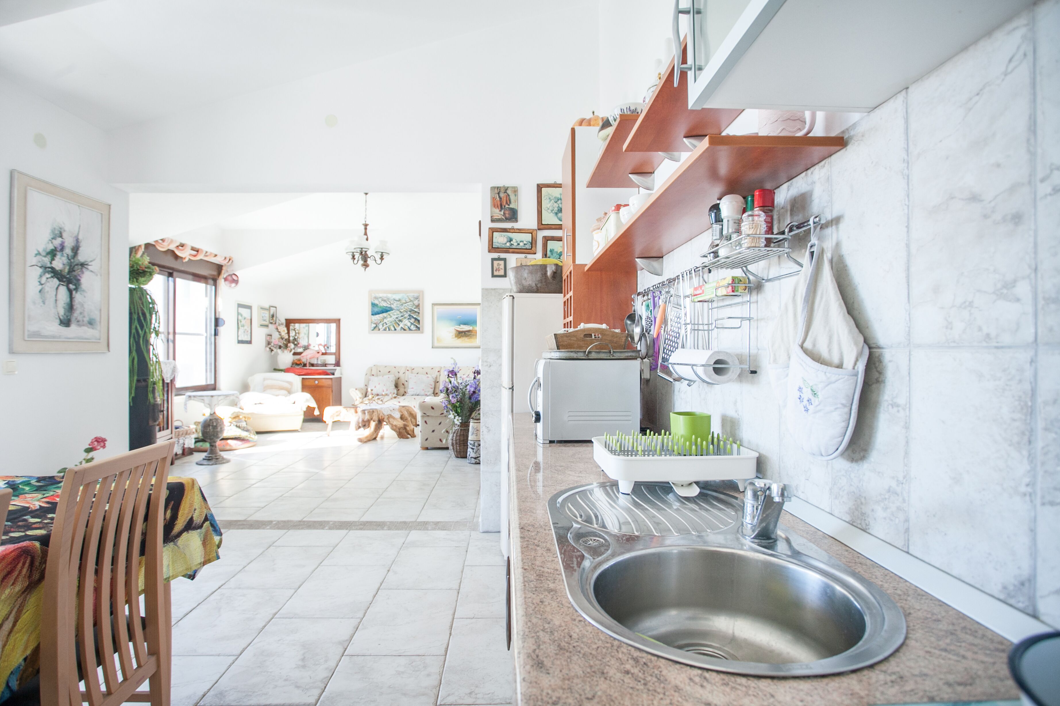 Una cucina con lavello, piano di lavoro e scaffalature. Si intravede, sullo sfondo, una zona pranzo con tavolo e sedie.