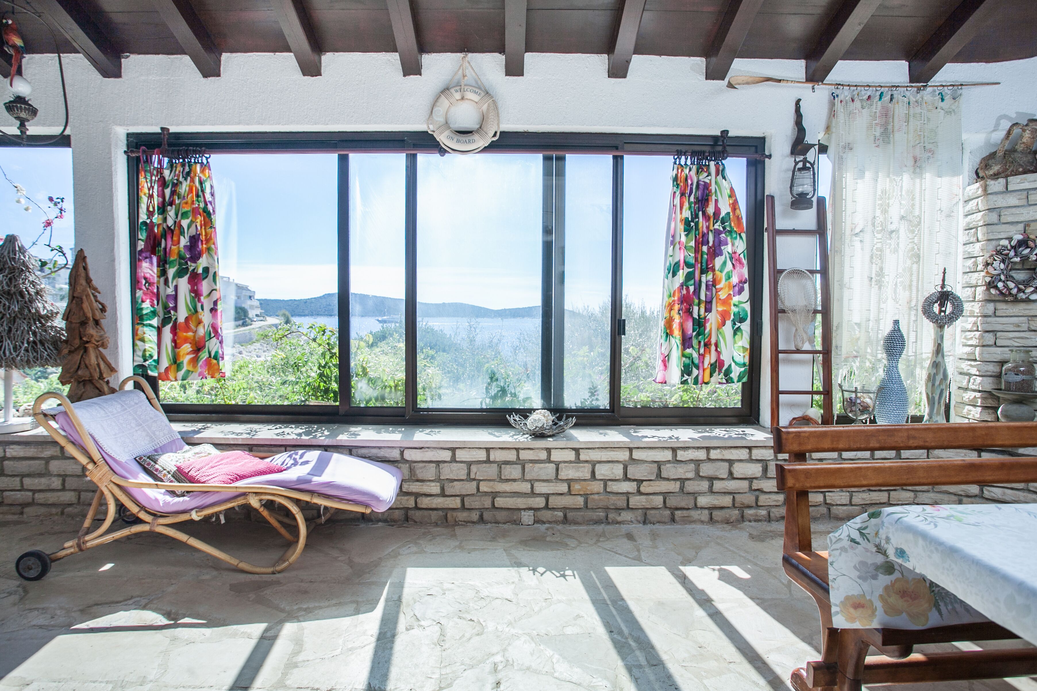 A room with a large window, a wicker chair, a wooden bed, and a stone wall.