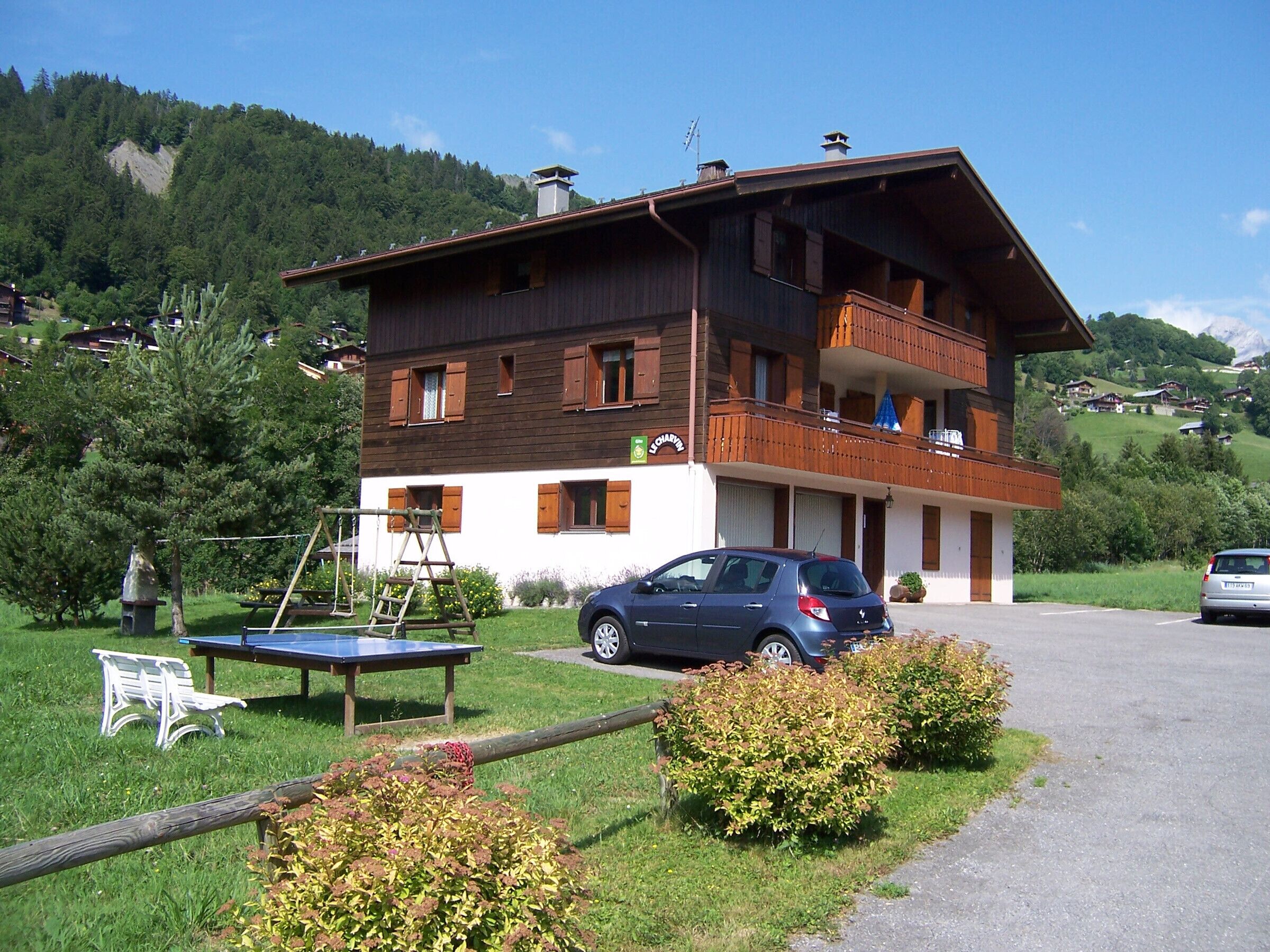 Ein zweistöckiges Holzhaus mit Balkon, ein Auto davor und ein kleiner Tisch mit einer Bank in der Nähe.