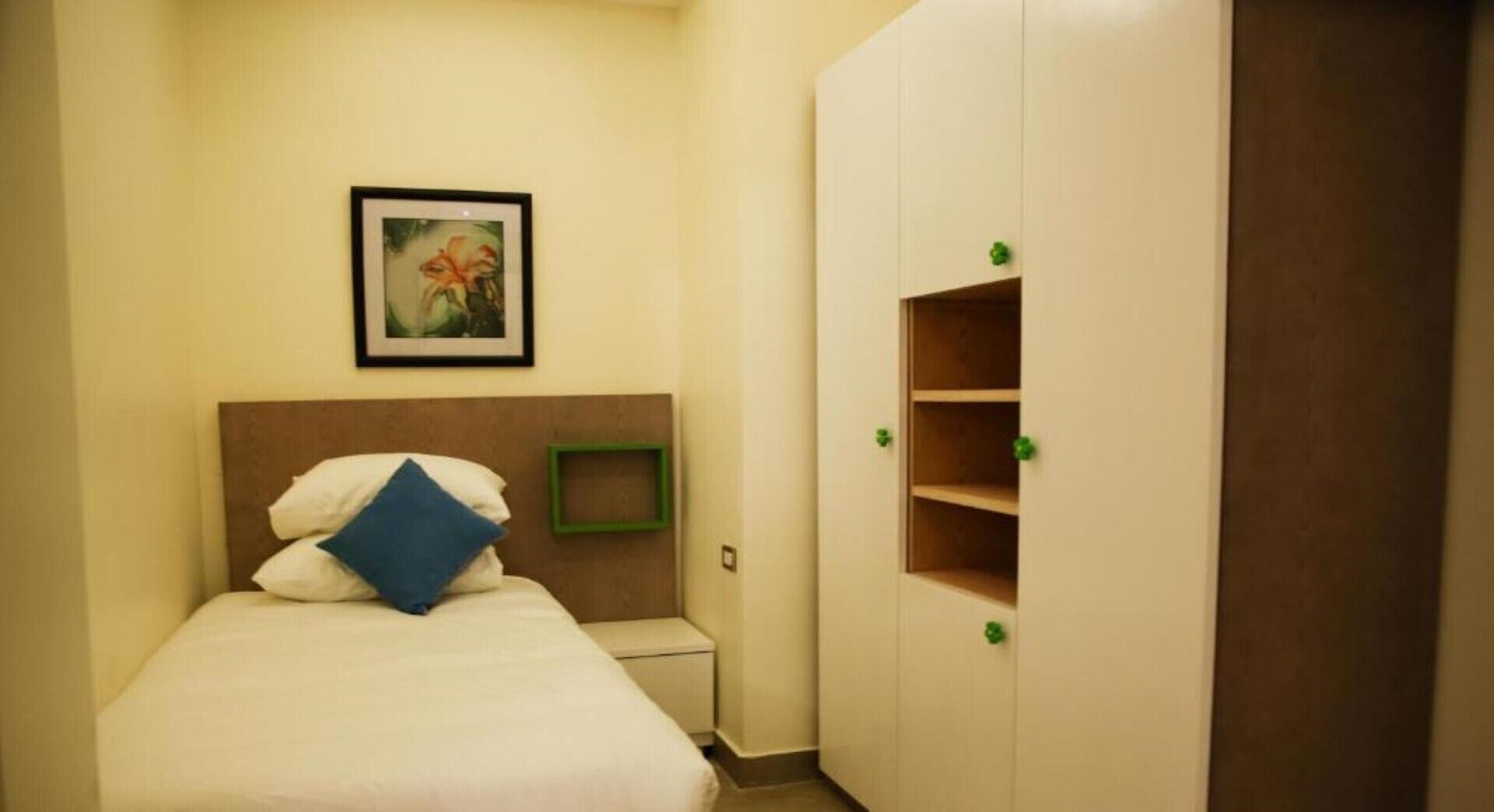 Habitación de hotel con cama, un cuadro en la pared, un armario con tiradores verdes y un pequeño estante verde.