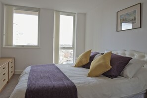 Een slaapkamer met een bed, twee ramen, een kaptafel en een lijst met een afbeelding aan de muur.