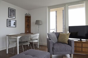 A modern living room with a grey sofa, a dining table with chairs, a TV, and a window with a view.