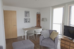 Een moderne woonkamer met een grijze bank, een televisie en een eethoek met een tafel en stoelen.
