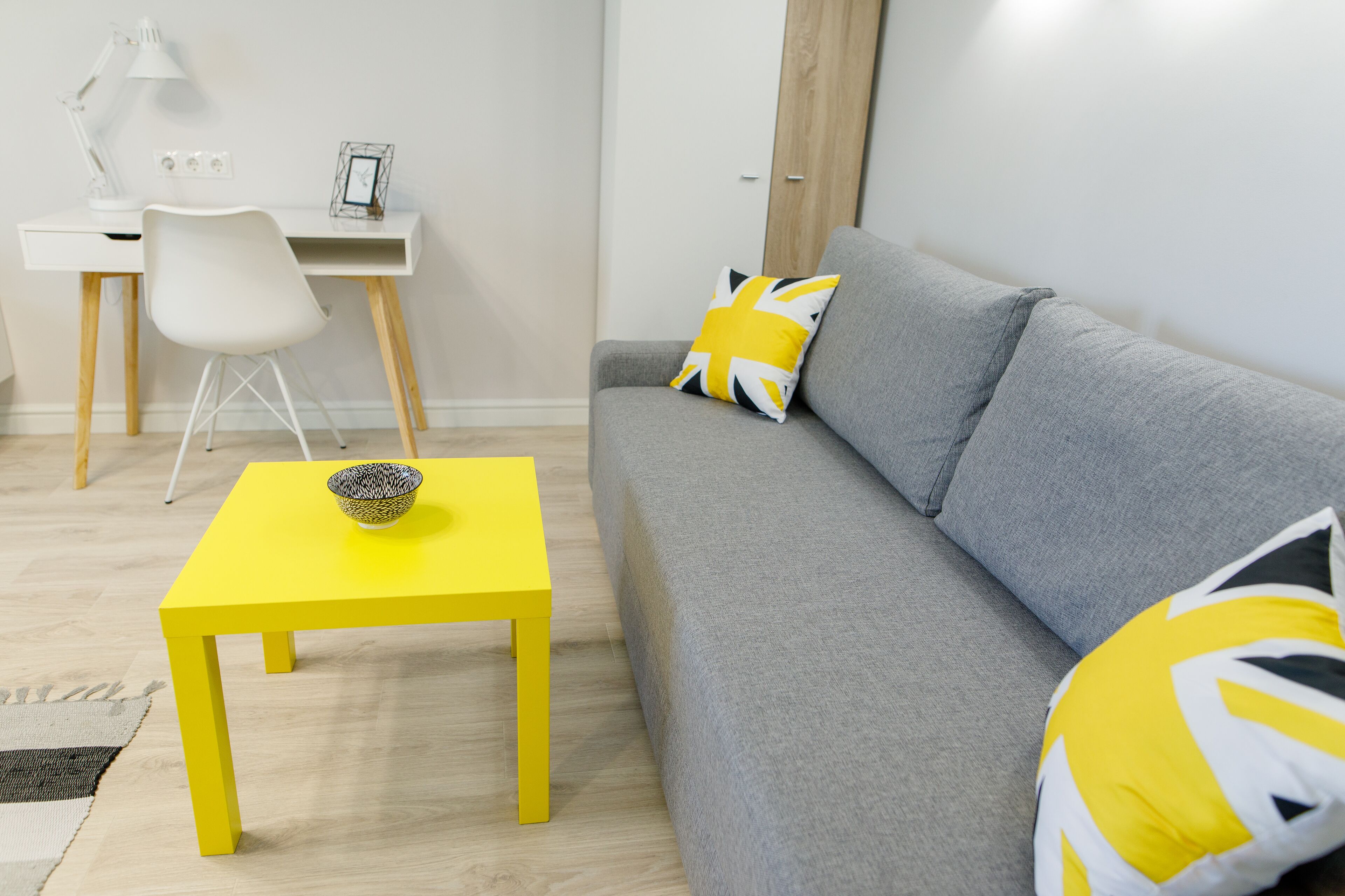 A modern living room with a grey sofa, a yellow table, and a white desk with a chair.