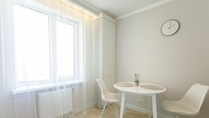 Una habitación con una mesa redonda blanca, dos sillas blancas, un reloj en la pared y una ventana con cortinas de encaje.