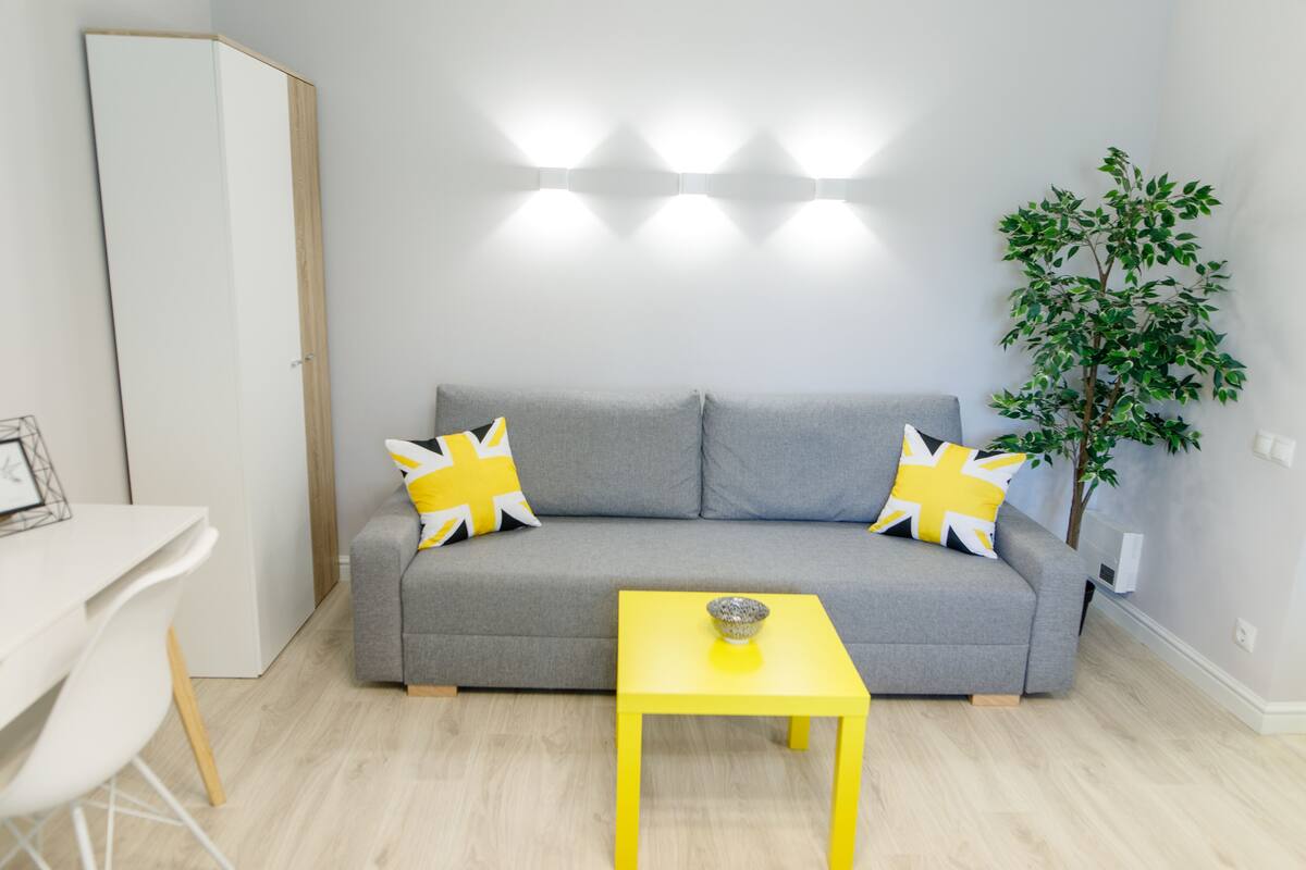 A modern living room with a grey sofa, yellow pillows, a yellow table, and a small plant.