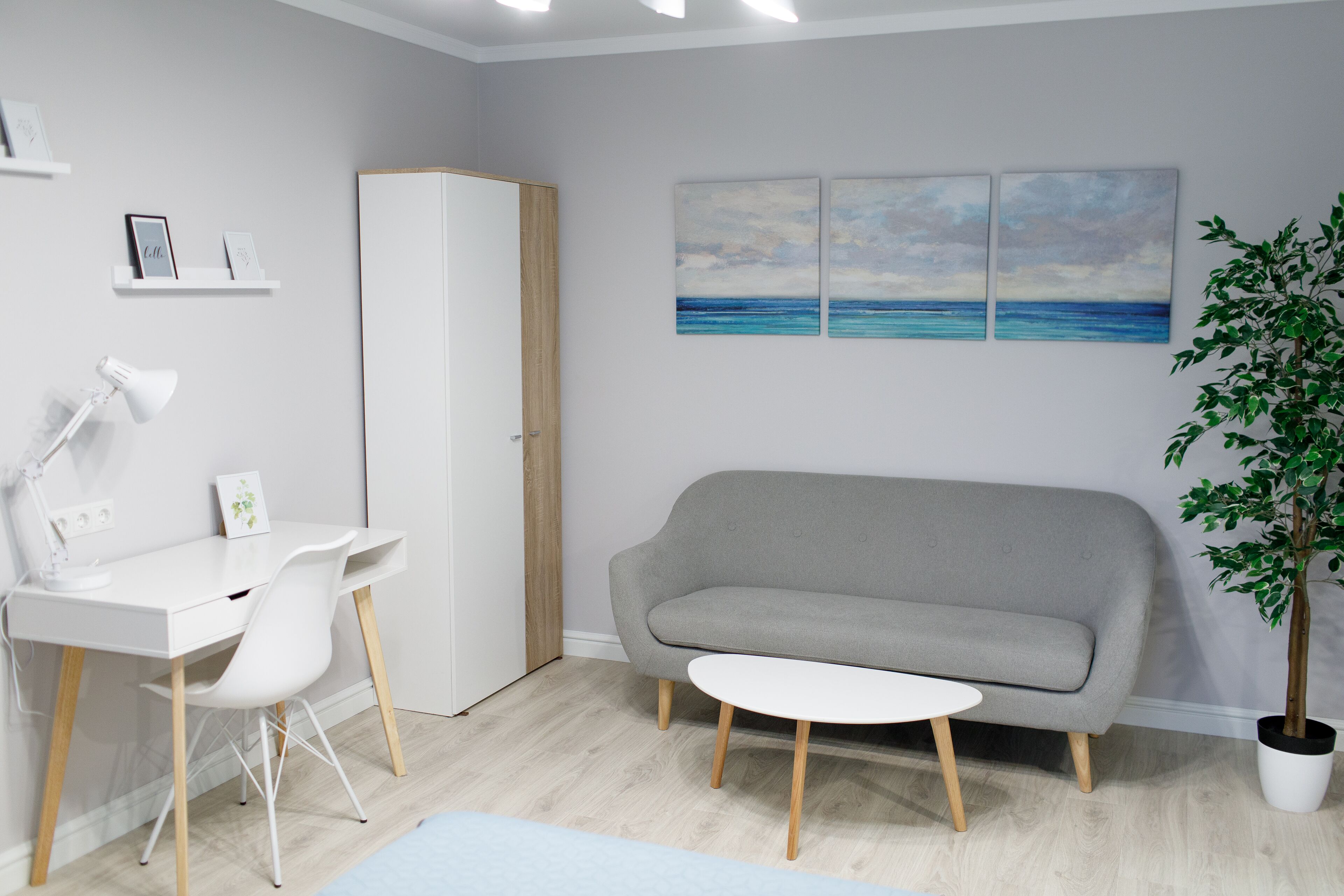 Un salón moderno con un escritorio blanco, un sofá gris, una pequeña mesa blanca y una planta en maceta.