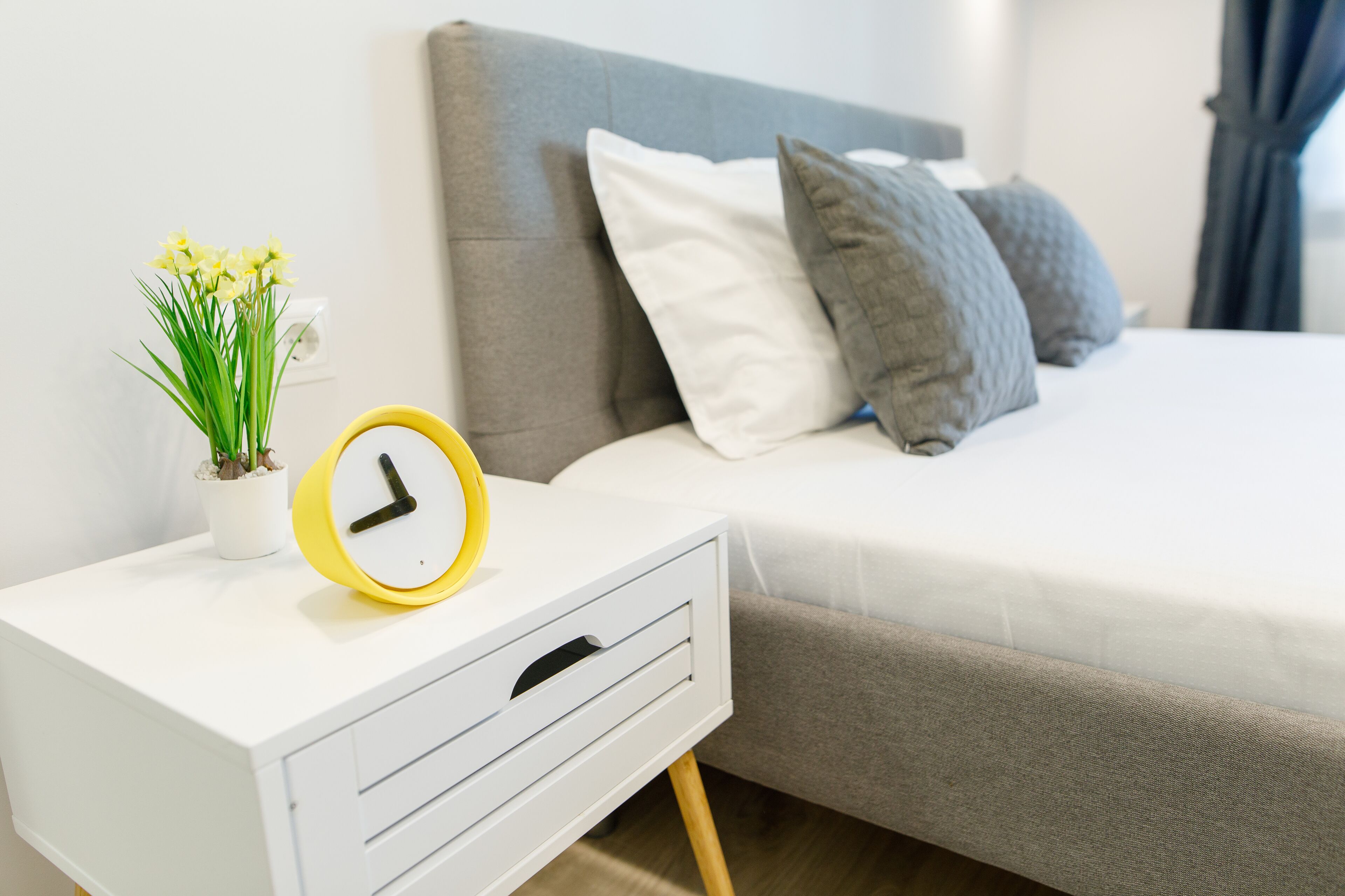 A neatly made bed with a grey headboard, a white bedside table with a yellow alarm clock, and a small potted plant.