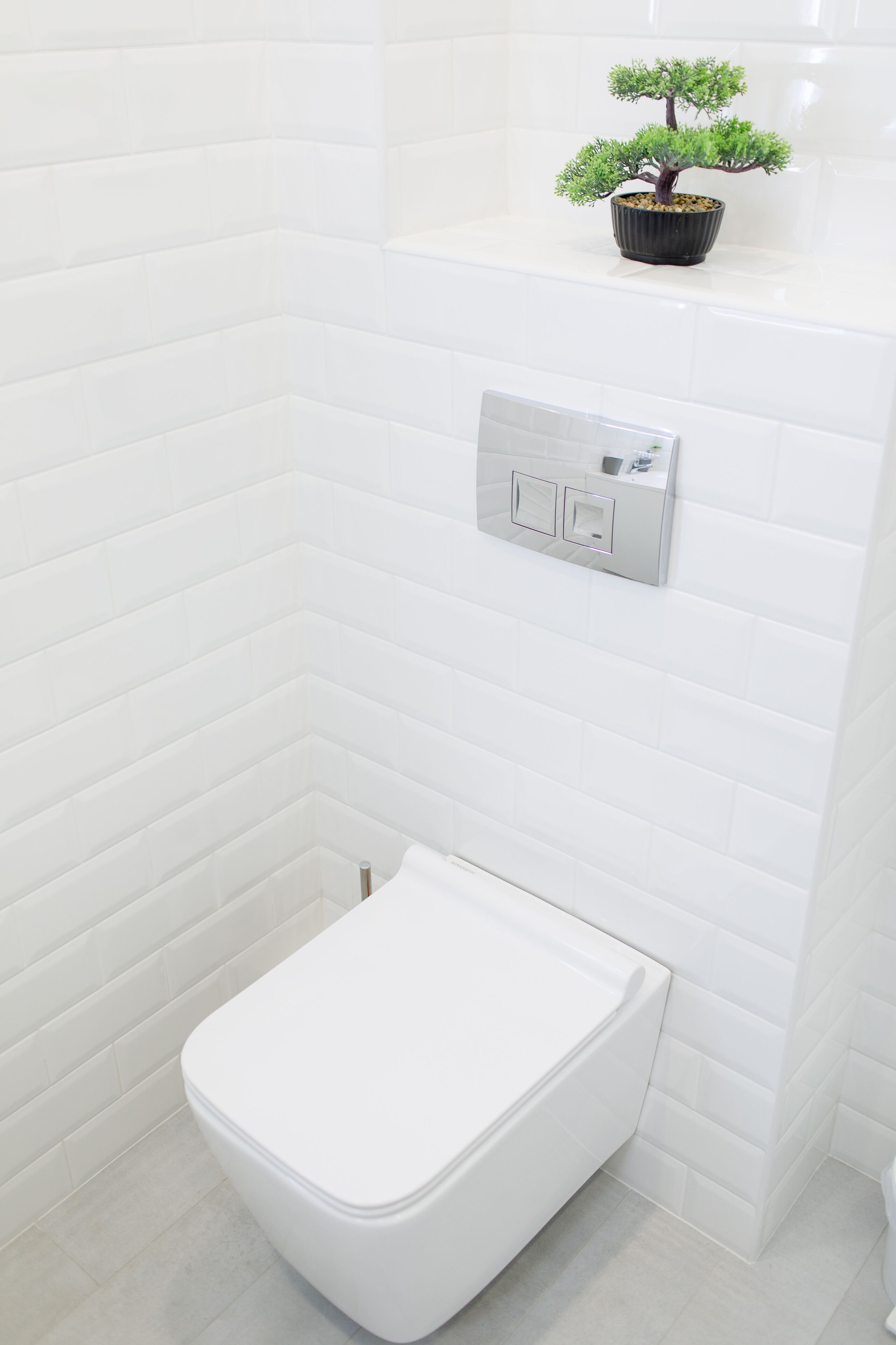 Une salle de bain moderne avec des carreaux de céramique blancs rectangulaires, des toilettes blanches et une petite plante en pot accrochée au mur.