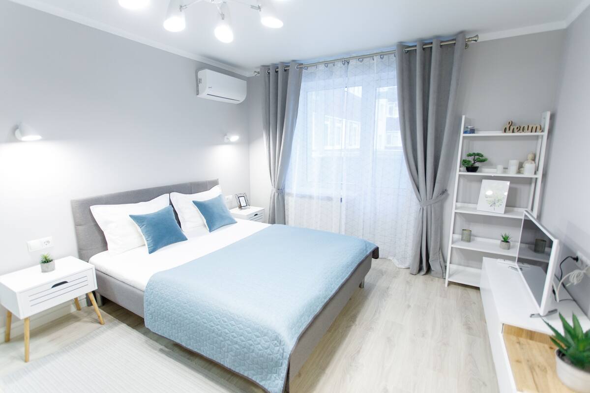 A modern bedroom with a bed, bedside tables, a nightstand, a shelf with decorative items, and a window with curtains.