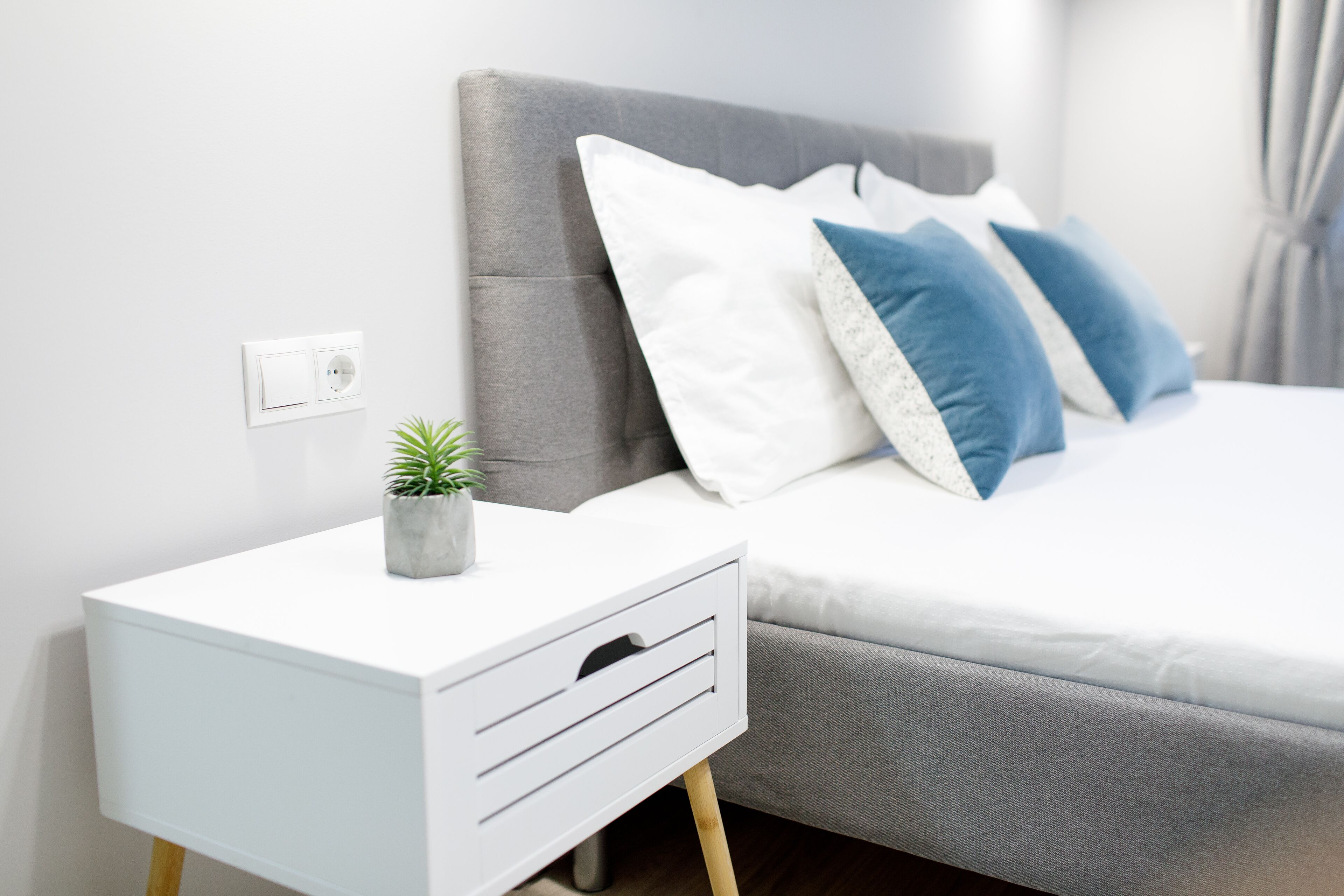 Un dormitorio moderno con cabecera tapizada gris, una mesita de noche blanca con cajones y una cama gris con almohadas blancas y azules.