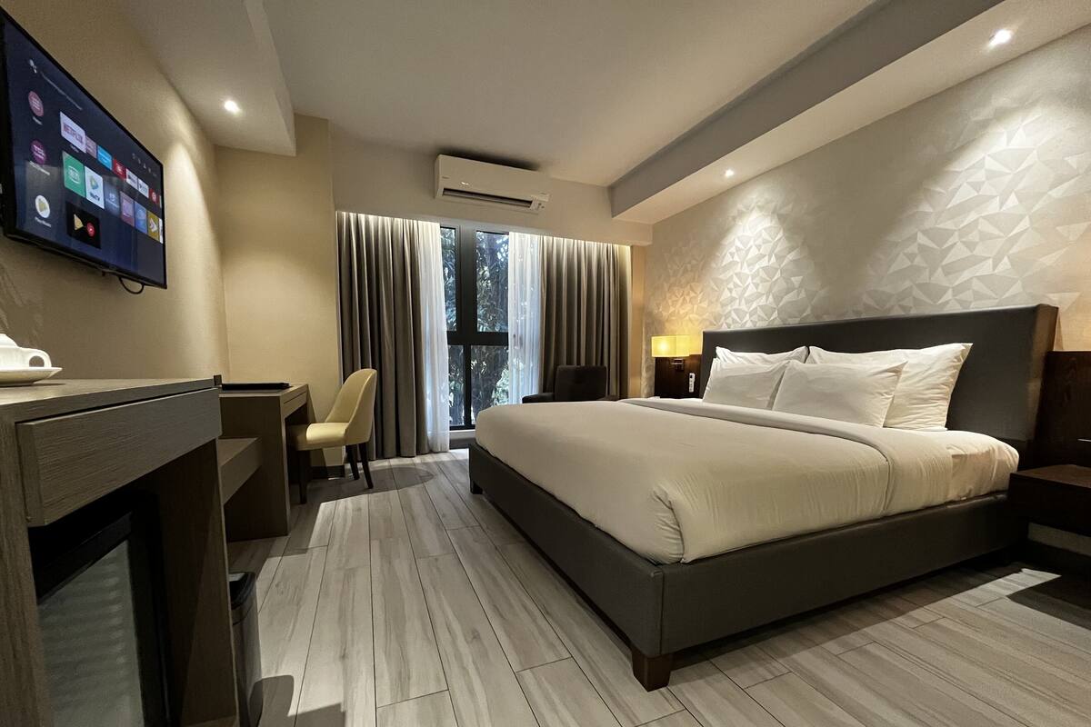 A hotel room with a bed, desk, chair, TV, and a window with curtains.