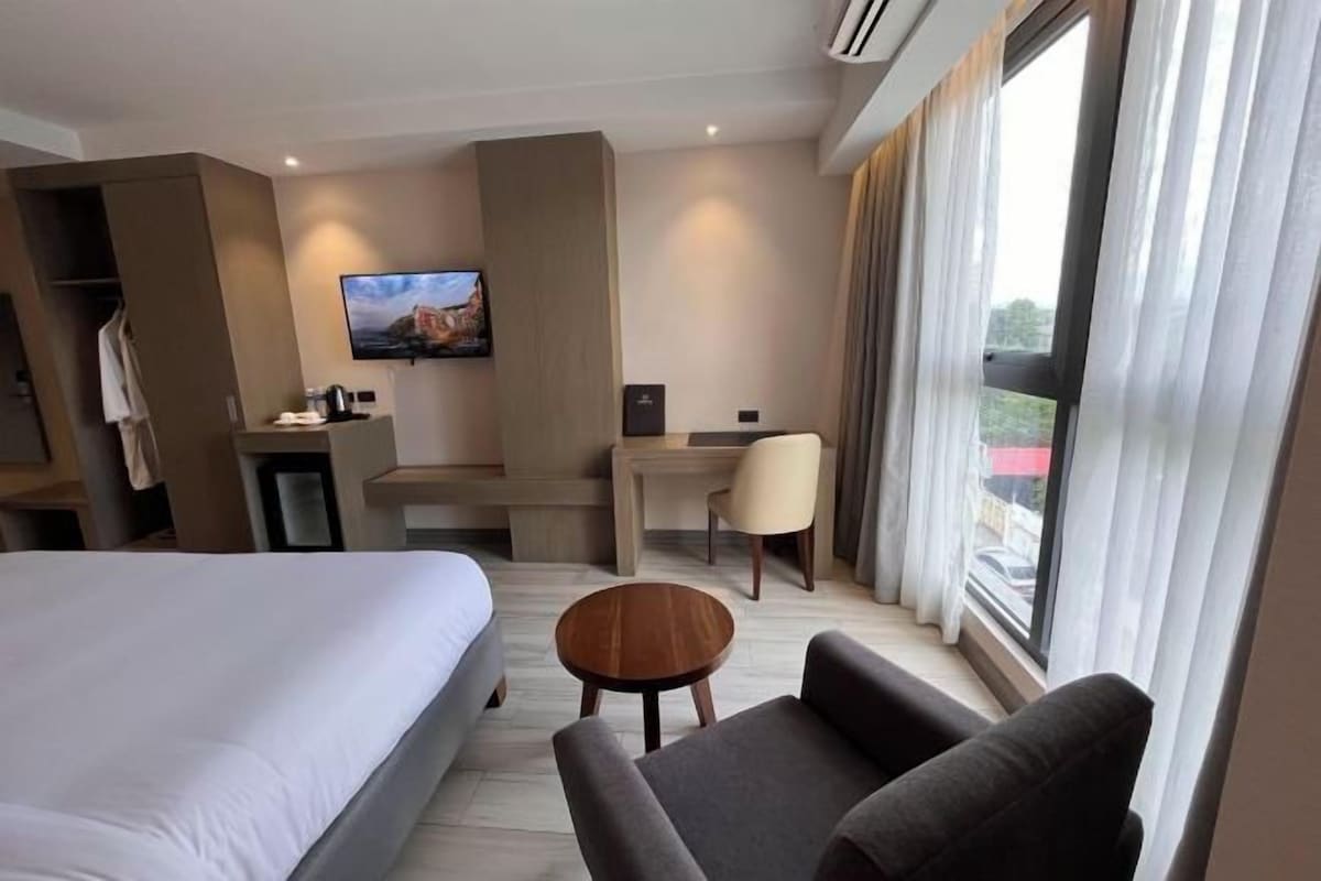 Quarto de hotel com cama, escrivaninha, cadeira, mesa pequena e televisão.