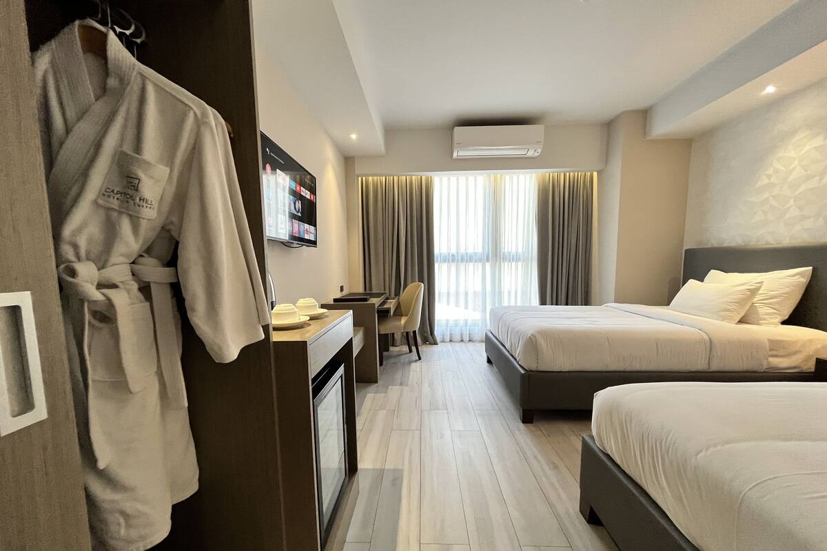 A hotel room with two beds, a desk with a chair, a robe hanging on a rack, and a TV mounted on the wall.