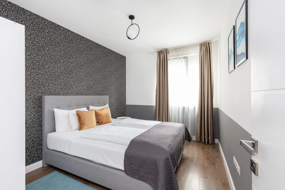 Una habitación de hotel moderna con una cama gris, ropa de cama blanca, una pared de acento gris con textura, una alfombra azul y una ventana con cortinas.