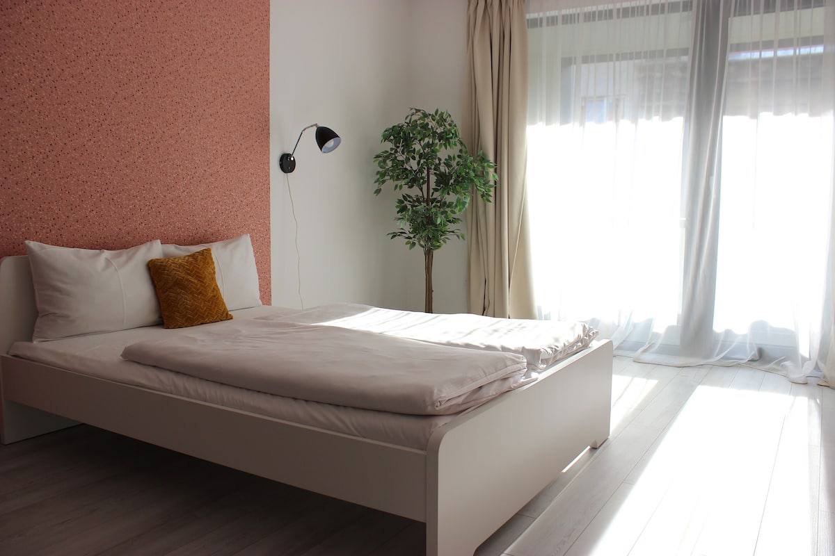 Una cama bien hecha con ropa de cama blanca y marrón, una lámpara de pared y una planta en maceta en una habitación luminosa con cortinas translúcidas.