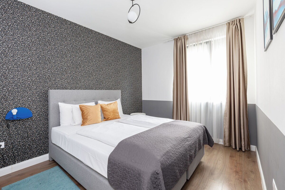 A neatly made bed with white and gray bedding, a textured wall behind it, and a window with beige curtains.