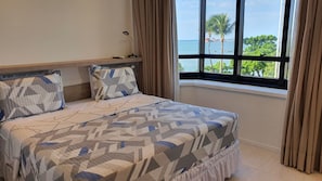 Una cama con colcha de diseño geométrico, una mesita de noche con lámpara y una ventana con vistas al mar y palmeras.