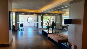 A spacious lobby with a modern design, featuring a seating area with cushioned benches, a large mirror, and a television mounted on the wall. - Apartamento Luxo em Hotel - Beira Mar Fortaleza (Fortaleza)