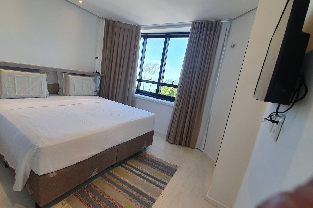 A bedroom with a large bed, a TV mounted on the wall, and a window with curtains.