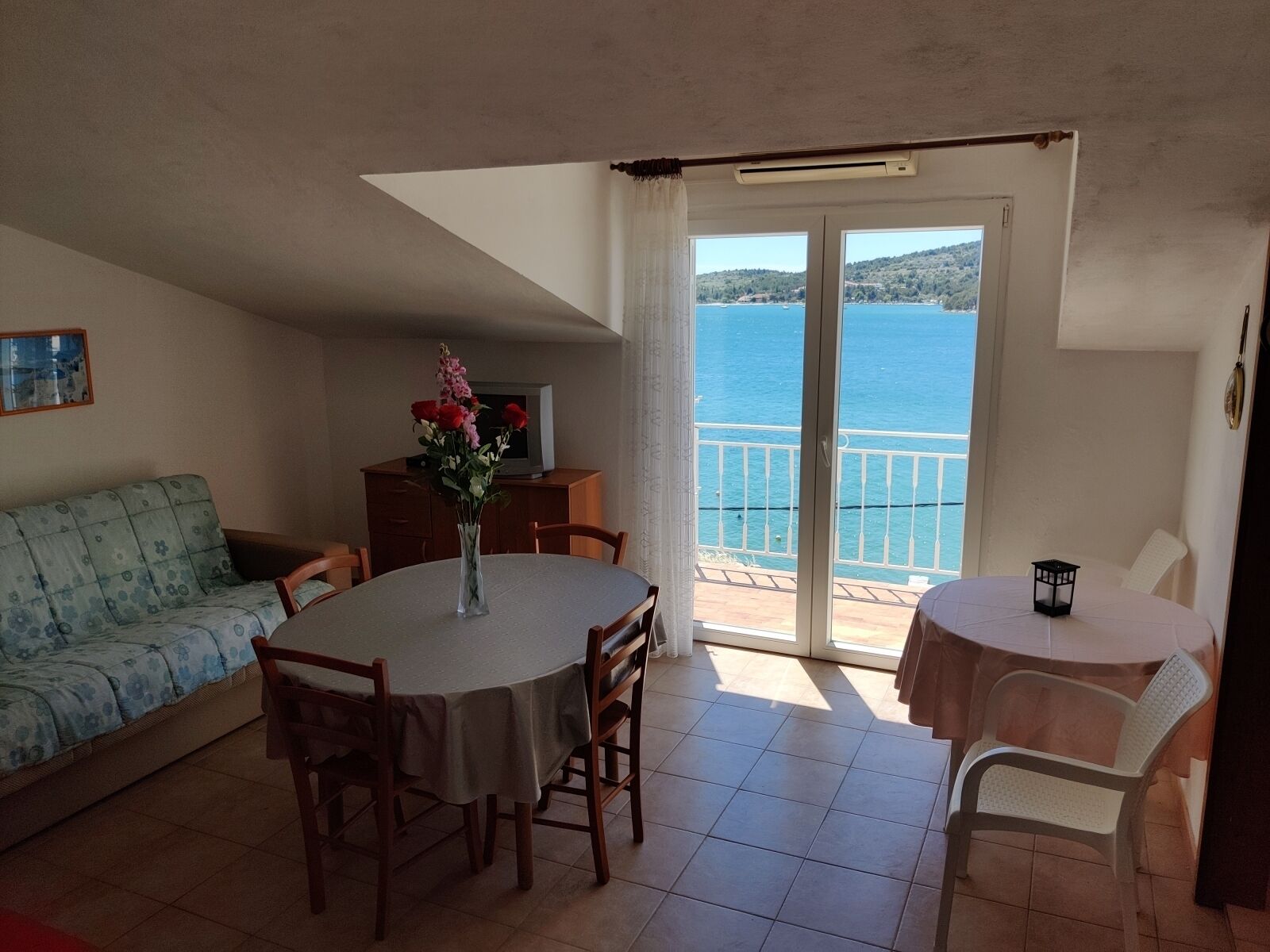A dining area with a table and chairs, a sofa, a TV, and a view of the sea through a sliding door.