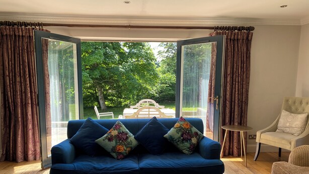 A living room with a blue sofa, a wooden coffee table, and a view of a garden through open curtains. - The Bridge House, Dunbar including hot tub (Dunbar)