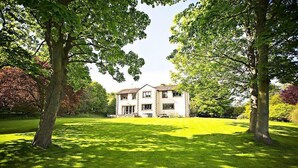 A two-story house with a front porch, surrounded by a well-maintained lawn and trees. - The Bridge House, Dunbar including hot tub (Dunbar)