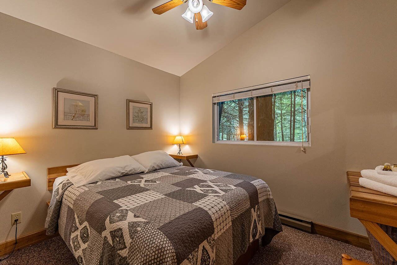 Une chambre à coucher avec un lit, deux lampes de chevet, une fenêtre donnant sur des arbres et un ventilateur de plafond.