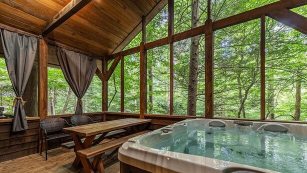 A wooden cabin with a hot tub, wooden benches, and large windows overlooking a forest.