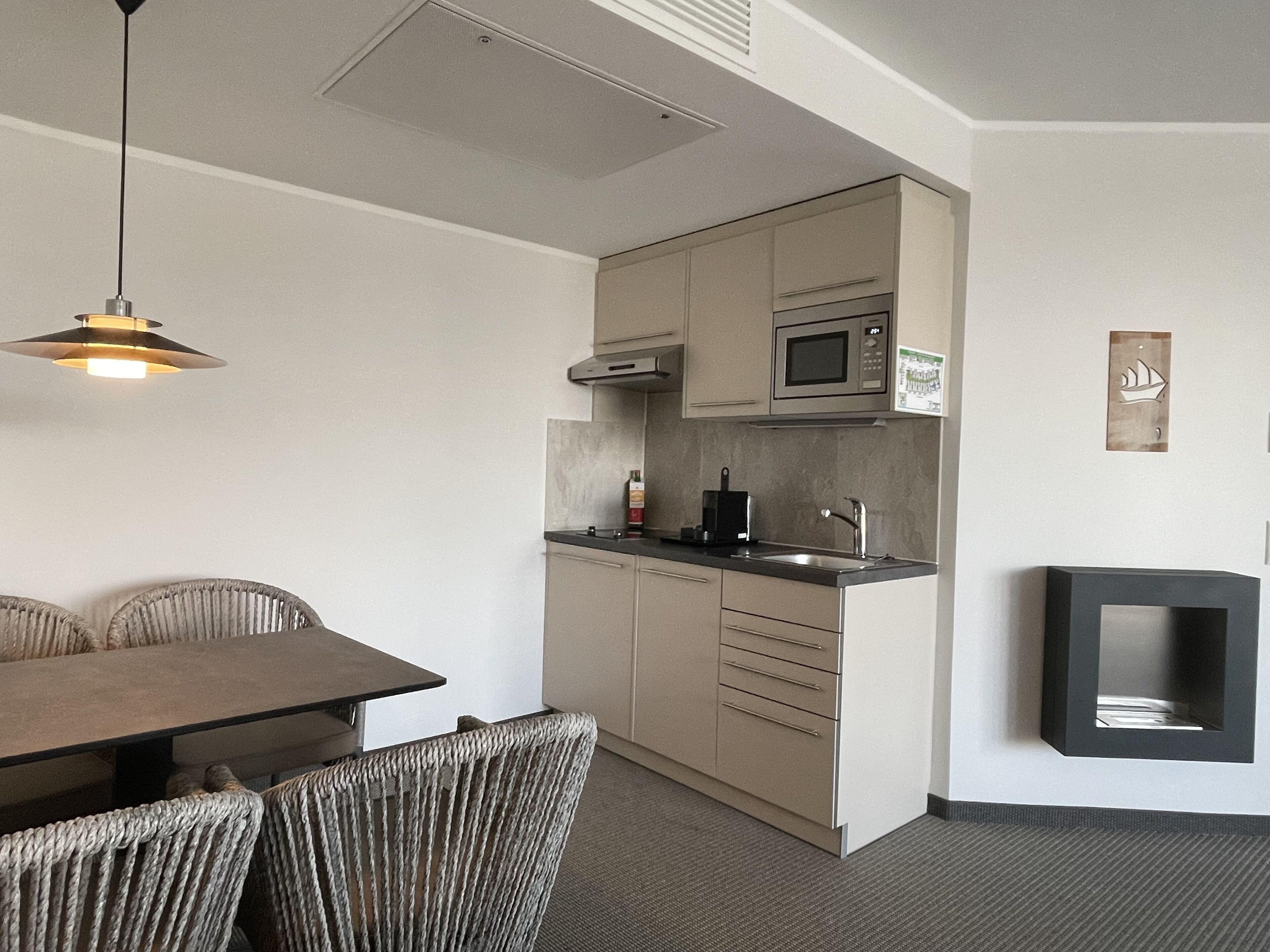 A compact kitchen with a microwave, sink, and cabinets. There is a dining table with chairs and a hanging light fixture.