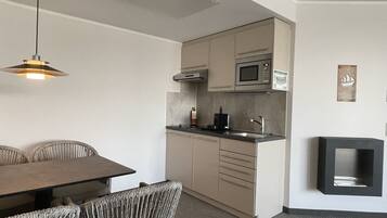 A compact kitchen with a microwave, sink, and cabinets. There is a dining table with chairs and a hanging light fixture.