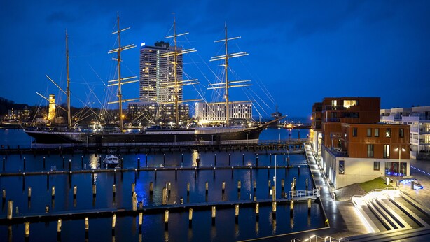 Un barco de vela histórico amarrado en un puerto deportivo con edificios modernos al fondo.