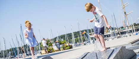 Children playing on a stone structure by a waterfront with sailboats in the background.