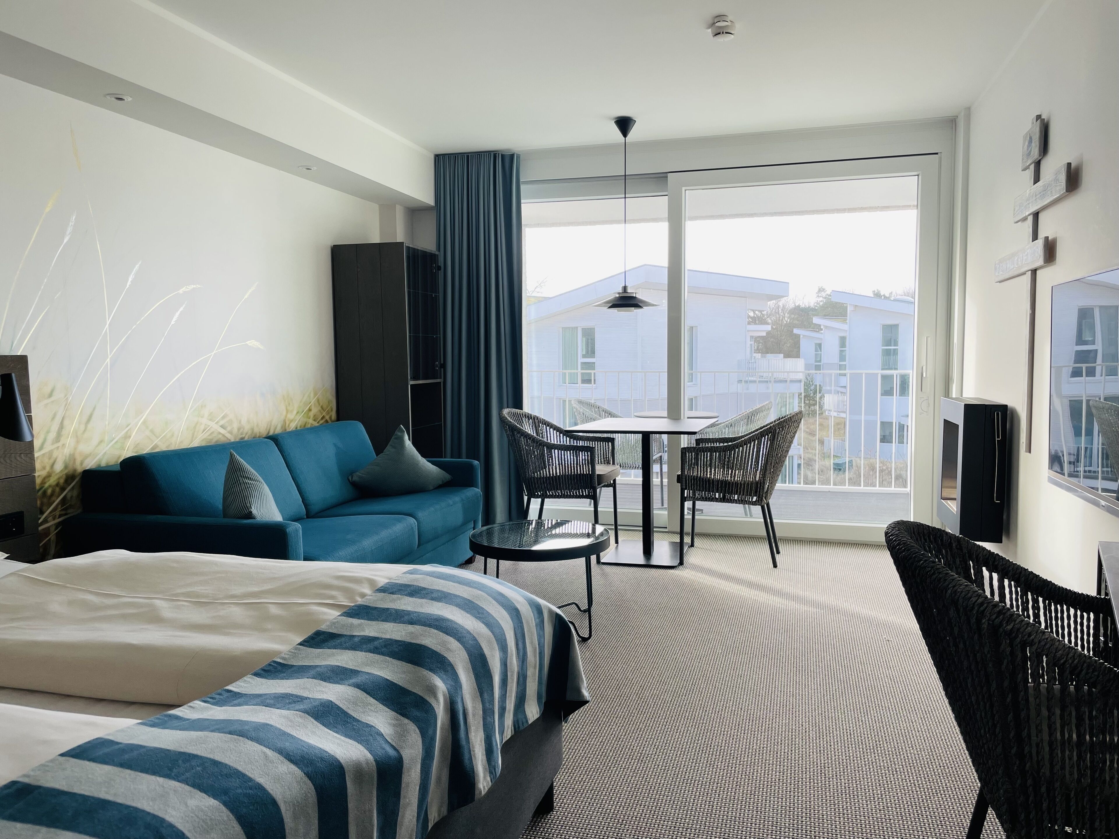 A modern hotel room with a large bed, a sofa, a dining table with chairs, a balcony with a view of buildings, and a ceiling fan.