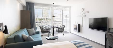 Moderni hotellihuone, jossa on suuri sänky, sohva, ruokapöytä tuoleineen, televisio ja parveke, jolta on näkymä rakennuksiin.